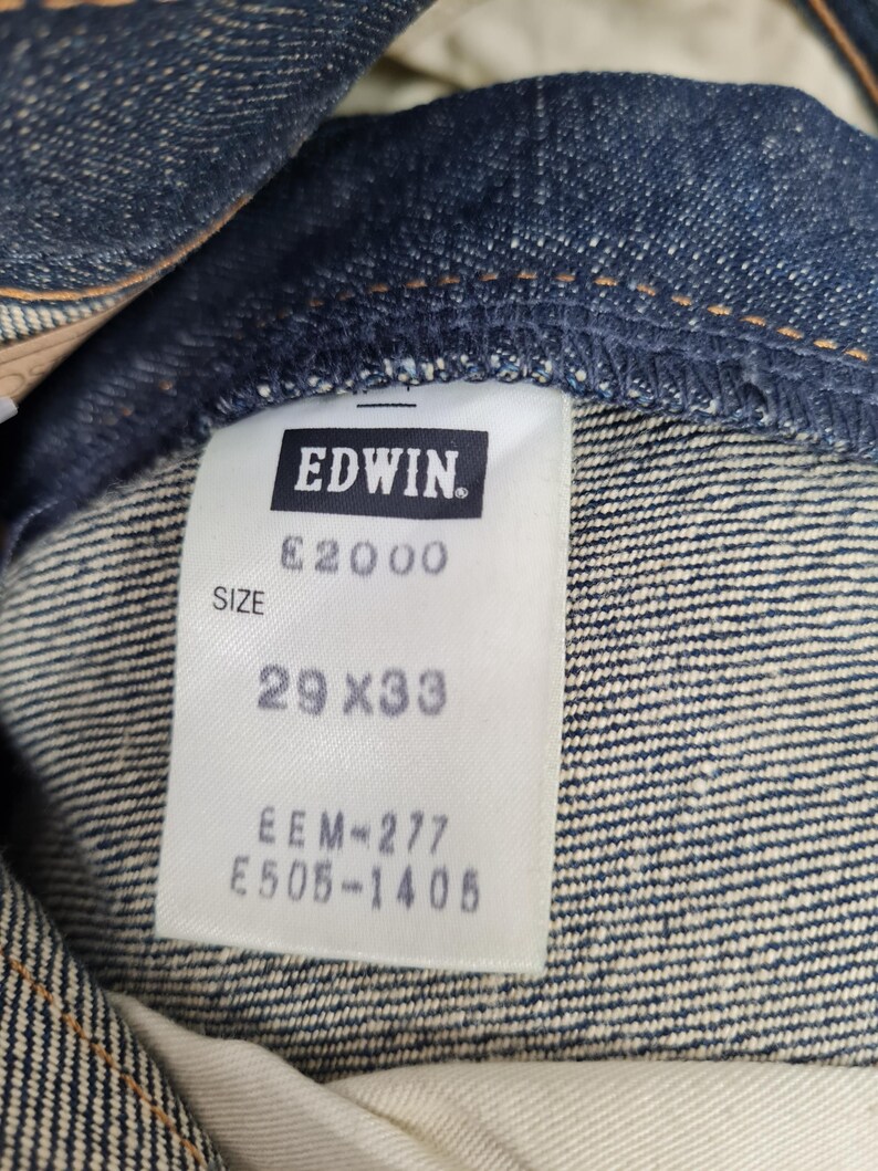 Op de afbeelding: Een wit label met zwarte tekst dat "EDWIN. E2000 SIZE 29 X 33 EEM=277 E505-1406" zegt, bevestigd aan een blauwe spijkerbroek.