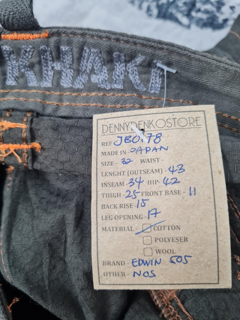 May include: Close-up of olive green denim pants with orange stitching. The word "KHAKI" is embroidered on the waistband. A tag reads "DENNYDENKOSTORE" and lists measurements: Outseam 43 inches, Inseam 34 inches, and Waist 32 inches. Brand is Edwin 505.