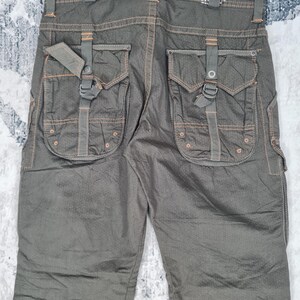 May include: Dark olive green cargo pants with multiple pockets and orange stitching accents. The pants have a textured appearance and feature a buckle and strap detail on the back pockets. The label reads "TYPE-505".