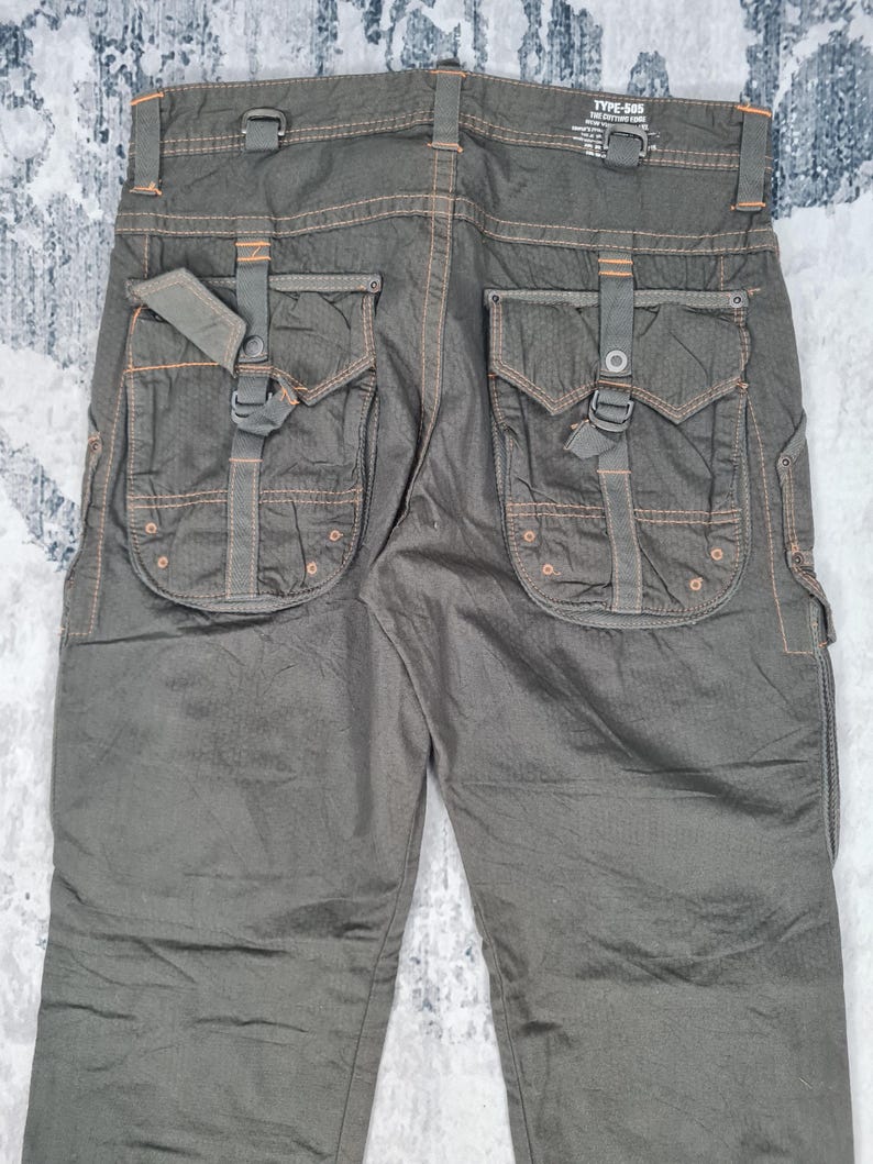 May include: Dark olive green cargo pants with multiple pockets and orange stitching accents. The pants have a textured appearance and feature a buckle and strap detail on the back pockets. The label reads "TYPE-505".