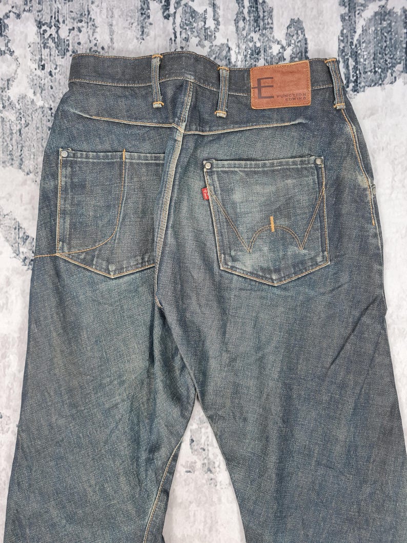 Op de afbeelding: Een paar donkerblauwe spijkerbroeken met een bruin leren patch op de achterkant van de tailleband met de tekst "Function Denim".
