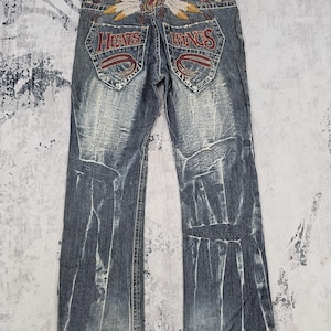 以下が含まれることがあります： 色あせた青色のデニムジーンズ。背面のポケットには、赤い刺繍で「HEARTS」と「WINGS」の文字と羽のデザインが施されています。ウエストバンドには革のパッチが見えます。