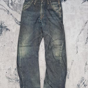 May include: A pair of blue denim jeans with a faded and distressed look. The jeans have a straight leg and a relaxed fit. The waistband has a red label with the word "Evisu" on it.