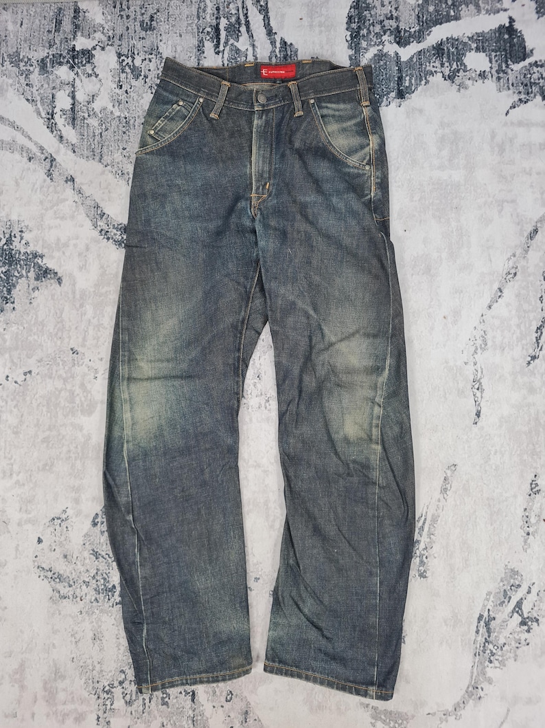Op de afbeelding: Een paar blauwe denim jeans met een vervaagde en verweerde look. De jeans heeft een rechte pijp en een ontspannen pasvorm. De tailleband heeft een rood label met het woord "Evisu" erop.