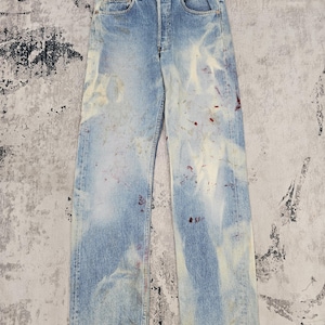 Puede incluir: Vaqueros de mezclilla azul claro con un lavado descolorido y salpicaduras de pintura. Los vaqueros tienen bragueta con botones y diseño clásico de cinco bolsillos. La mezclilla tiene un aspecto desgastado con detalles en blanco y rojo.