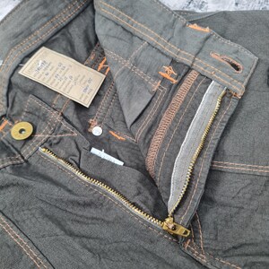 May include: Close-up of olive green pants with a textured honeycomb pattern. The pants feature orange stitching, a brass zipper, and a button closure. A tag with text is attached to the waistband.