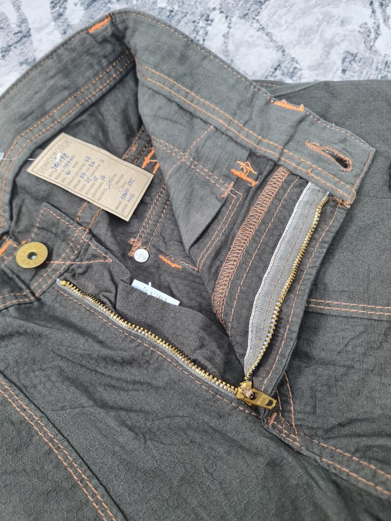 May include: Close-up of olive green pants with a textured honeycomb pattern. The pants feature orange stitching, a brass zipper, and a button closure. A tag with text is attached to the waistband.