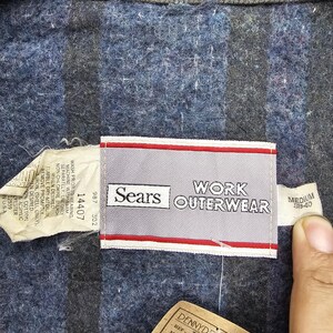 Puede incluir: Una chaqueta de trabajo azul con una etiqueta blanca y roja que dice "Sears Work Outerwear" y "Medium 38-40". La etiqueta tambi&eacute;n incluye el n&uacute;mero de referencia "J0077" y la talla "M".