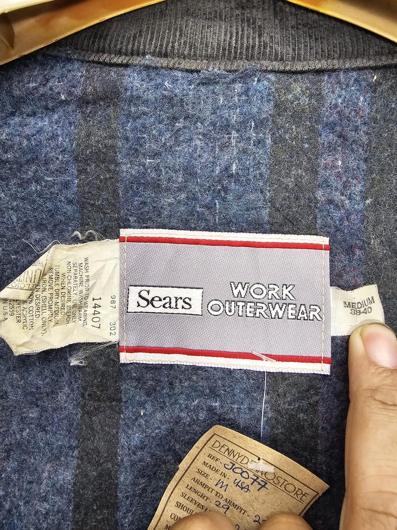 Puede incluir: Una chaqueta de trabajo azul con una etiqueta blanca y roja que dice "Sears Work Outerwear" y "Medium 38-40". La etiqueta tambi&eacute;n incluye el n&uacute;mero de referencia "J0077" y la talla "M".