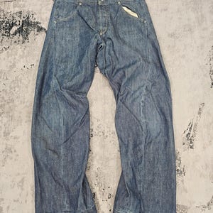 Puede incluir: Vaqueros de mezclilla azules con bragueta de botones y diseño de cinco bolsillos. Los vaqueros tienen un corte relajado y un dobladillo ligeramente desgastado. Los vaqueros están planos sobre una superficie texturizada.