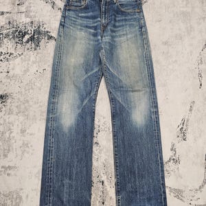 Puede incluir: Unos vaqueros de mezclilla azul con un lavado medio que presenta un desgaste intencional en los muslos y las rodillas. Los pantalones tienen un diseño clásico de cinco bolsillos, cierre de botones y corte recto con costuras visibles.