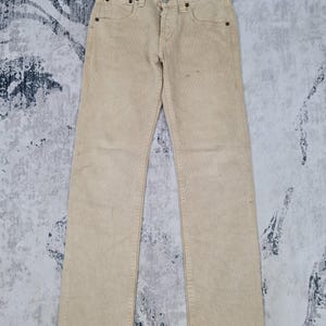 Könnte beinhalten: Eine hellbeige gerade Jeanshose. Die Jeans hat ein klassisches Fünf-Taschen-Design und einen Knopfverschluss.