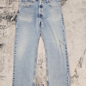 May include: Light blue denim jeans with a classic five-pocket design. The jeans have a straight leg cut and show signs of wear, including fading and slight fraying at the hem. The jeans are laid flat on a textured surface.