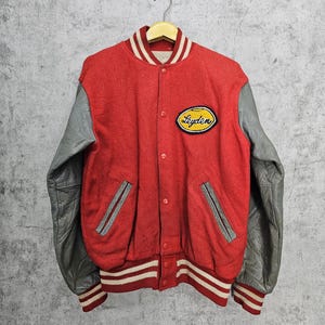 Puede incluir: Chaqueta de béisbol de lana roja con mangas de cuero gris y cuello y puños a rayas blanco y rojo. La chaqueta tiene un parche amarillo con la palabra "Leyden" en el pecho izquierdo.