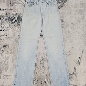 Puede incluir: Vaqueros de mezclilla azul claro con bragueta de botones y diseño clásico de cinco bolsillos. Los vaqueros tienen un corte de pierna recta y muestran un sutil desgaste. La prenda está tendida sobre una superficie texturizada.