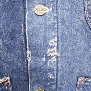 Puede incluir: Primer plano de una chaqueta vaquera azul con dos botones de lat&oacute;n que dicen "SEARS". Los botones est&aacute;n cosidos en la parte delantera de la chaqueta.