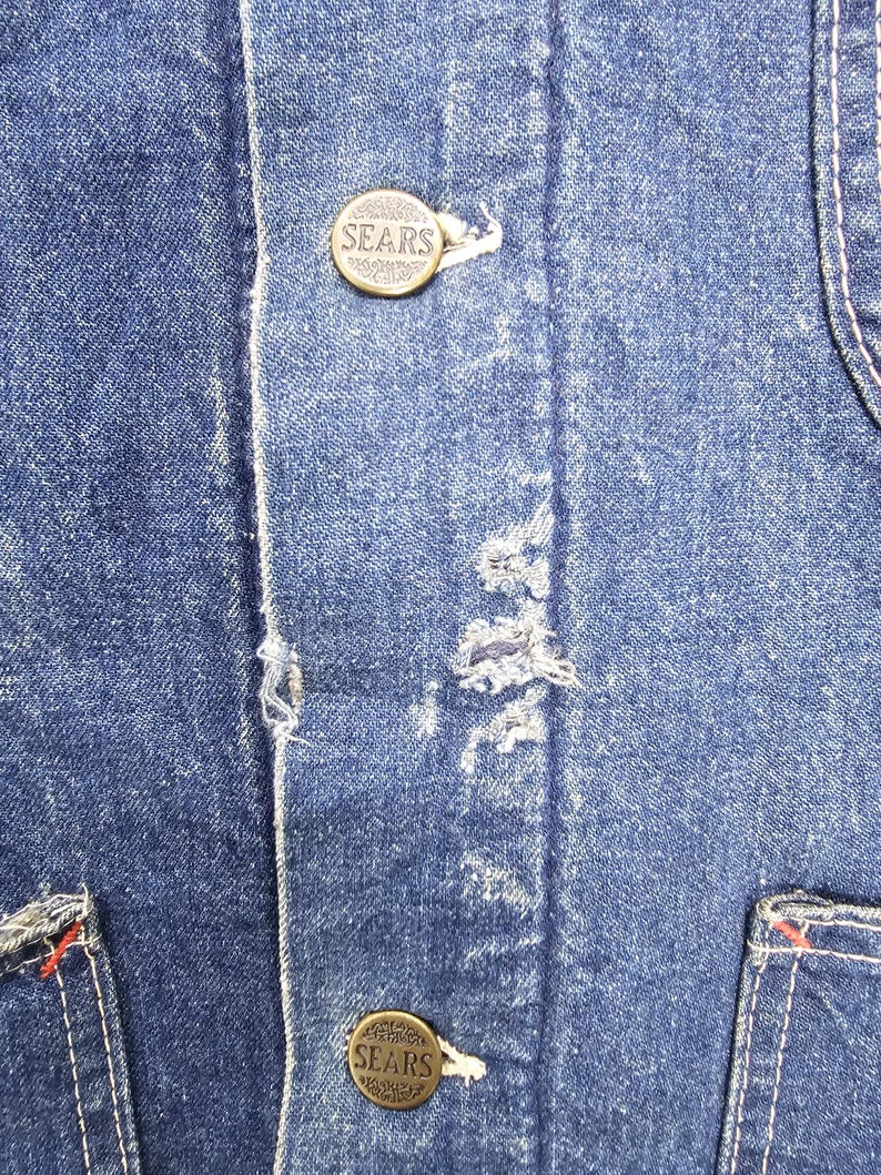 Puede incluir: Primer plano de una chaqueta vaquera azul con dos botones de lat&oacute;n que dicen "SEARS". Los botones est&aacute;n cosidos en la parte delantera de la chaqueta.