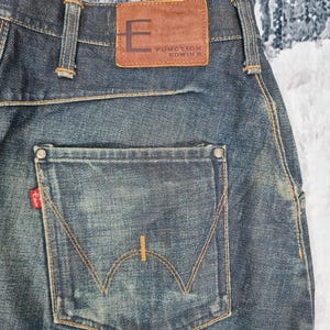 Op de afbeelding: Een paar blauwe denim jeans met een bruin leren patch op de achterkant van de tailleband met de tekst "Function Edwin". De achterzak heeft een opvallend gebogen stikselontwerp.