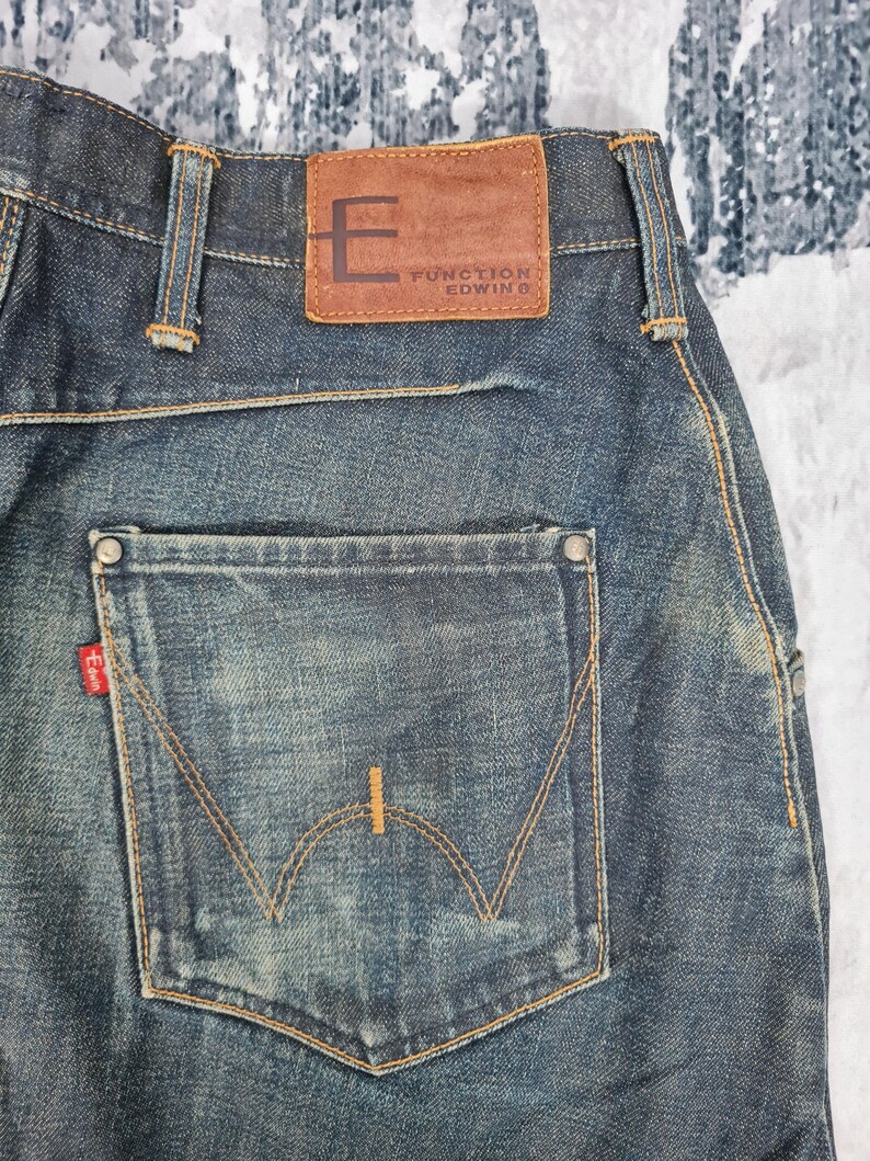 Op de afbeelding: Een paar blauwe denim jeans met een bruin leren patch op de achterkant van de tailleband met de tekst "Function Edwin". De achterzak heeft een opvallend gebogen stikselontwerp.