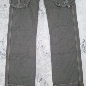 May include: Dark olive green cargo pants with multiple pockets and decorative stitching. The pants have a textured fabric and feature large pockets with buckle closures. The stitching is a contrasting brown color.