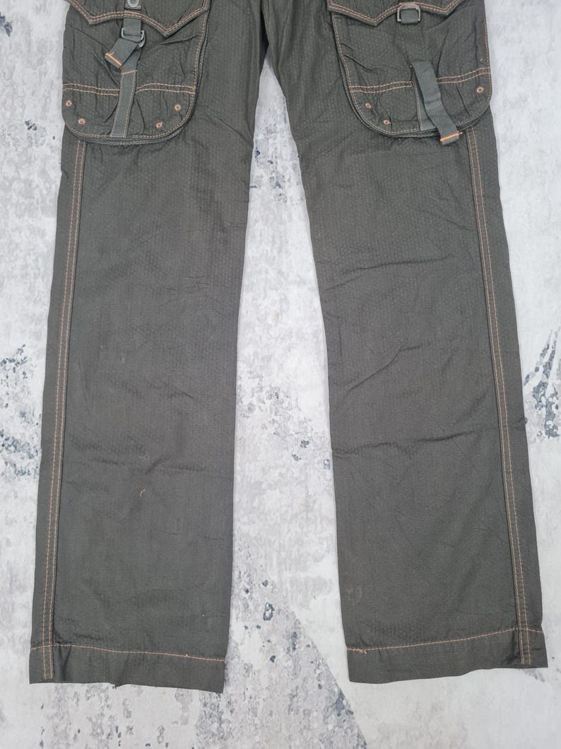 May include: Dark olive green cargo pants with multiple pockets and decorative stitching. The pants have a textured fabric and feature large pockets with buckle closures. The stitching is a contrasting brown color.