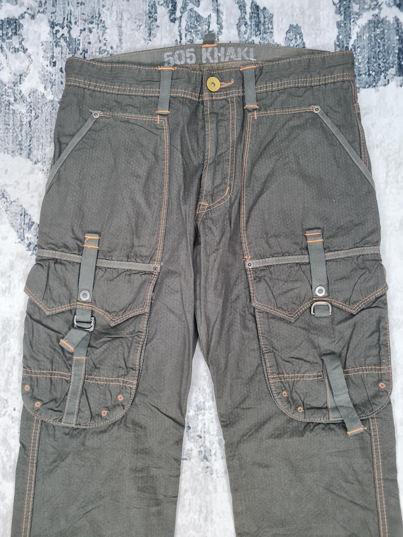 May include: Khaki-colored cargo pants with multiple pockets and orange stitching. The pants have a button closure and belt loops. The text "505 KHAKI" is visible on the waistband.