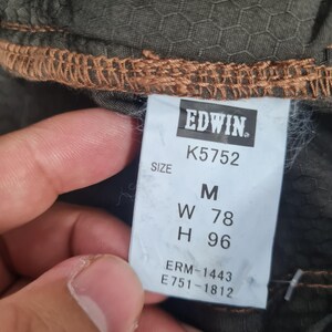 May include: Close-up of a tag from Edwin clothing. The tag is white with black text, displaying the brand name, size, and product codes. The size is M, with measurements W 78 and H 96. The fabric is olive green with a honeycomb pattern.