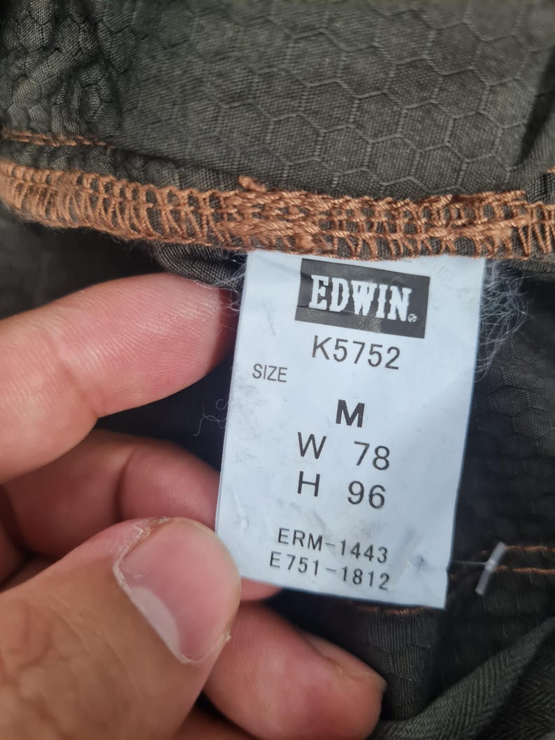 May include: Close-up of a tag from Edwin clothing. The tag is white with black text, displaying the brand name, size, and product codes. The size is M, with measurements W 78 and H 96. The fabric is olive green with a honeycomb pattern.
