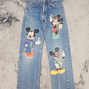Puede incluir: Vaqueros de mezclilla azul con tres estampados gráficos distintos de Mickey Mouse con diferentes atuendos. Los pantalones presentan un diseño clásico de cinco bolsillos con cierre de botones y remaches metálicos, corte de pierna recta e ilustraciones coloridas.