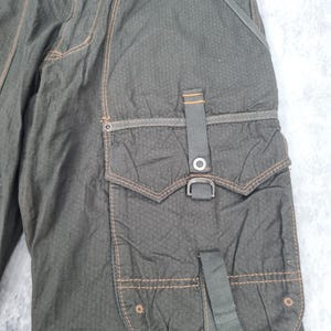 May include: Close-up of olive green cargo pants with multiple pockets and orange stitching. The pants have a textured fabric and a metal buckle detail on the side pocket. The design includes a button closure and belt loops.