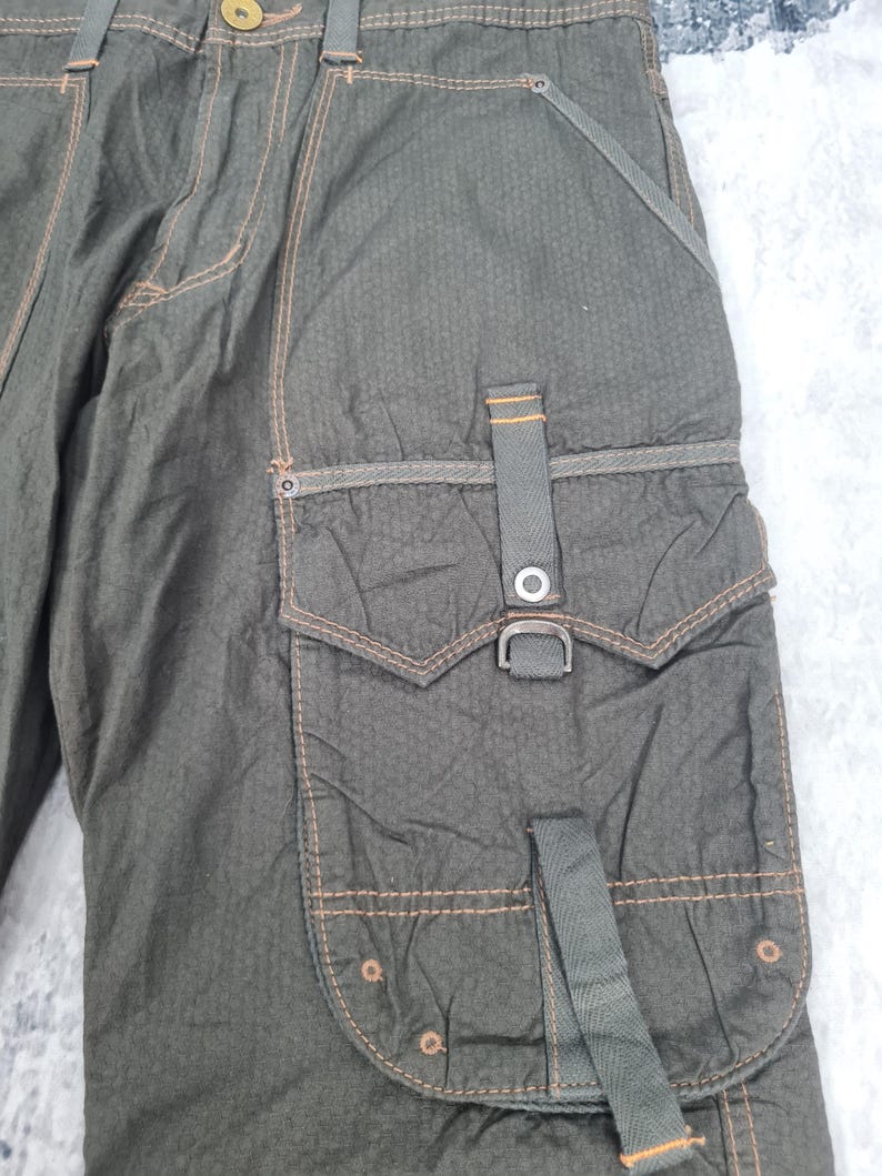 May include: Close-up of olive green cargo pants with multiple pockets and orange stitching. The pants have a textured fabric and a metal buckle detail on the side pocket. The design includes a button closure and belt loops.