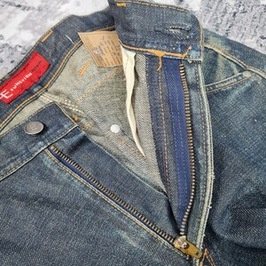 Op de afbeelding: Close-up van een paar blauwe denim jeans met een ritsvlieg. De jeans heeft een vervaagde en verweerde look. De tailleband heeft een rood label met de tekst "E FUNCTION DOWN".
