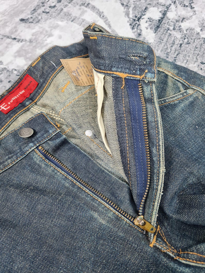 Op de afbeelding: Close-up van een paar blauwe denim jeans met een ritsvlieg. De jeans heeft een vervaagde en verweerde look. De tailleband heeft een rood label met de tekst "E FUNCTION DOWN".