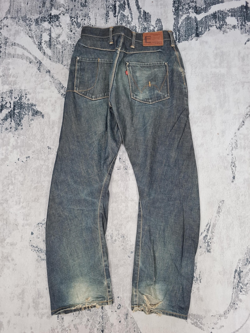 Op de afbeelding: Een paar blauwe denim jeans met distressed details en een rood leren patch op de achterkant van de tailleband. De jeans heeft een rechte pijp en een faded wash.