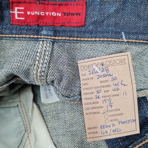 Op de afbeelding: Een blauw denim jean label met witte stiksels. Het label heeft de tekst "Function Edwin" in rood en de tekst "Denny Denko Store" in zwart. Het label bevat ook details over de jeans, zoals de maat, taille, inseam en materiaal.
