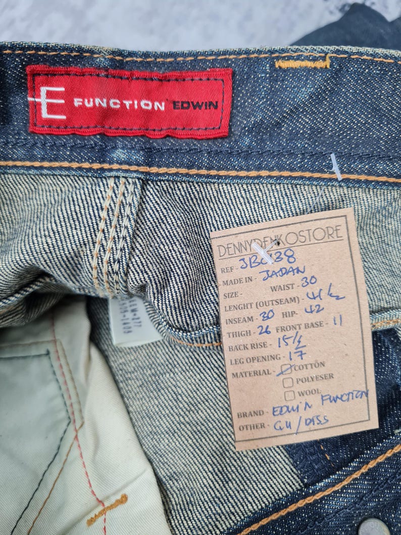Op de afbeelding: Een blauw denim jean label met witte stiksels. Het label heeft de tekst "Function Edwin" in rood en de tekst "Denny Denko Store" in zwart. Het label bevat ook details over de jeans, zoals de maat, taille, inseam en materiaal.