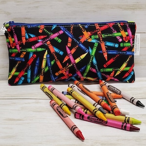 Peut inclure: Une pochette noire à fermeture éclair avec un imprimé de crayons de couleur. La pochette est remplie de crayons de couleur.