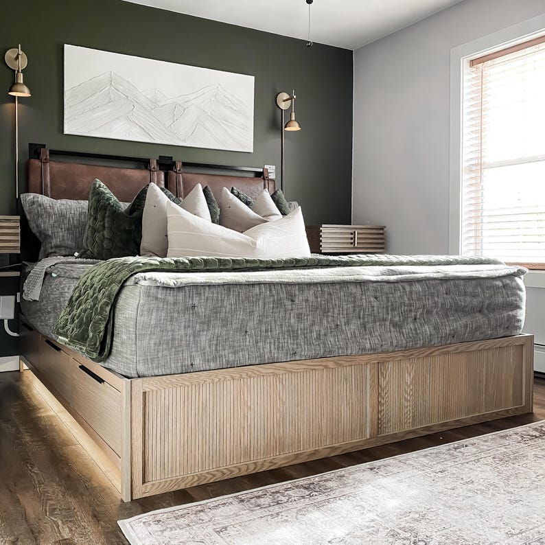 K&ouml;nnte beinhalten: Ein graues, gepolstertes Bettgestell mit einem Kopfteil und einem Fu&szlig;teil aus Holz. Das Bett hat eine gr&uuml;ne Wolldecke und wei&szlig;e Kissen. Das Bett steht in einem Schlafzimmer mit einer gr&uuml;nen Wand und einer wei&szlig;en Decke.