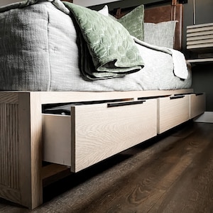 K&ouml;nnte beinhalten: Ein Holzbettgestell mit drei Schubladen, die von unten herausgezogen werden. Die Schubladen sind aus hellem Holz und haben schwarze Griffe. Das Bett ist mit einer hellgrauen und gr&uuml;nen Decke bedeckt.
