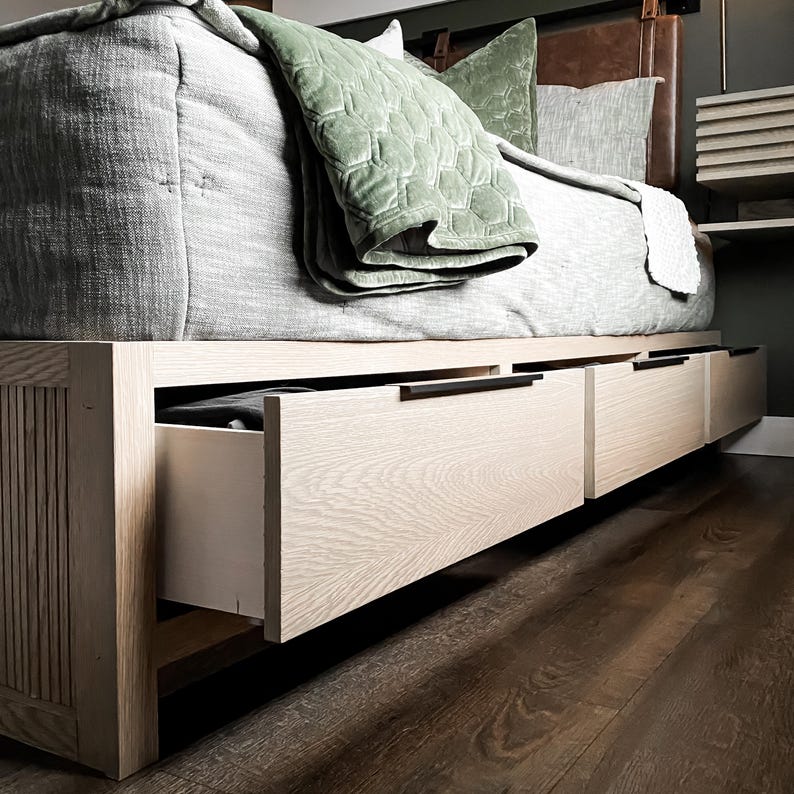 K&ouml;nnte beinhalten: Ein Holzbettgestell mit drei Schubladen, die von unten herausgezogen werden. Die Schubladen sind aus hellem Holz und haben schwarze Griffe. Das Bett ist mit einer hellgrauen und gr&uuml;nen Decke bedeckt.