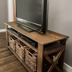 May include: A dark brown wooden TV stand with an X-shaped design on the bottom. The stand has three shelves, with two wicker baskets on the bottom shelves and a DVD player on the middle shelf. A flat screen TV is on top of the stand.