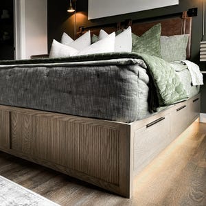 K&ouml;nnte beinhalten: Ein Holzbettgestell mit einem Lattenrost und Schubladen. Das Bett ist mit einem grauen gesteppten Bettbezug und gr&uuml;nen Samtkissen ausgestattet.
