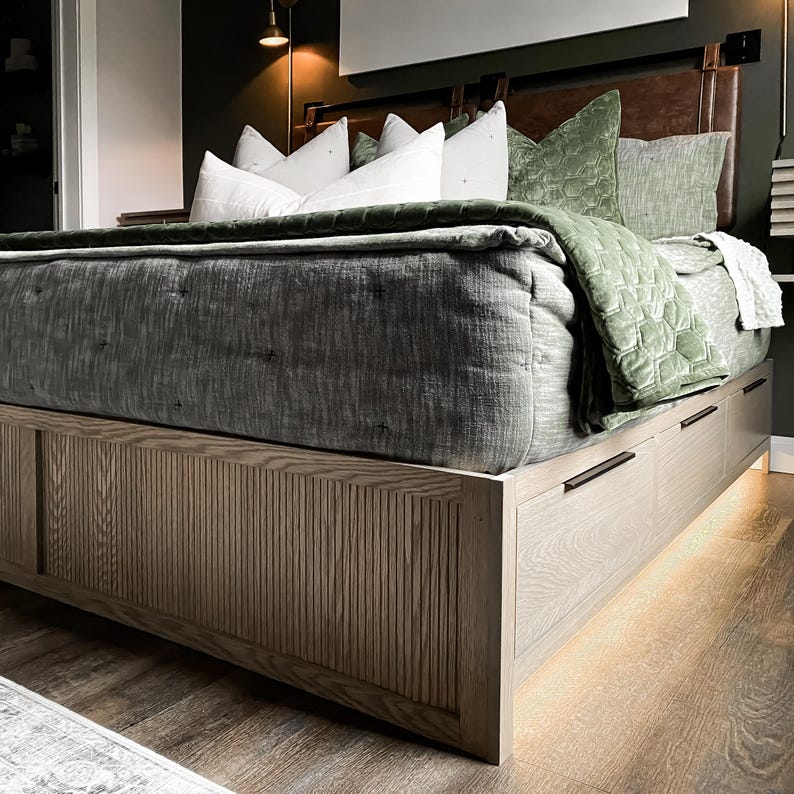 K&ouml;nnte beinhalten: Ein Holzbettgestell mit einem Lattenrost und Schubladen. Das Bett ist mit einem grauen gesteppten Bettbezug und gr&uuml;nen Samtkissen ausgestattet.