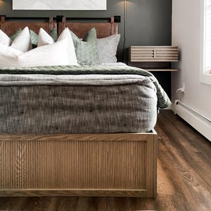K&ouml;nnte beinhalten: Ein Holzbettgestell mit einem Lattenrost am Kopf- und Fu&szlig;ende. Das Bett ist mit einer grauen Bettw&auml;sche aus Leinen und gr&uuml;nen Samtkissen bezogen. Das Bett steht auf einem Holzboden.
