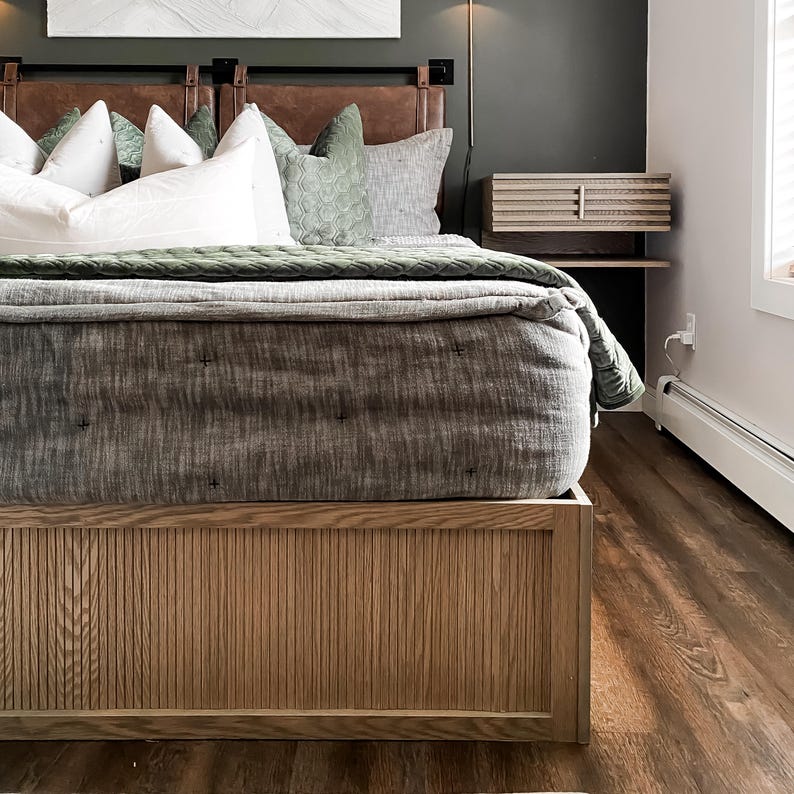 K&ouml;nnte beinhalten: Ein Holzbettgestell mit einem Lattenrost am Kopf- und Fu&szlig;ende. Das Bett ist mit einer grauen Bettw&auml;sche aus Leinen und gr&uuml;nen Samtkissen bezogen. Das Bett steht auf einem Holzboden.