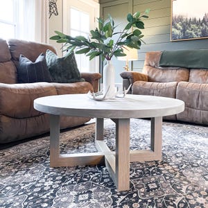 Puede incluir: Una mesa de centro redonda de hormigón con una tapa de color gris claro y una base de madera natural. La mesa está situada sobre una alfombra con estampado en blanco y negro en una sala de estar con dos sofás de cuero marrón.