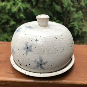 May include: White ceramic butter dish with a domed lid and a speckled glaze. The lid has a knob on top and the dish has a base. The dish is decorated with blue flower designs.