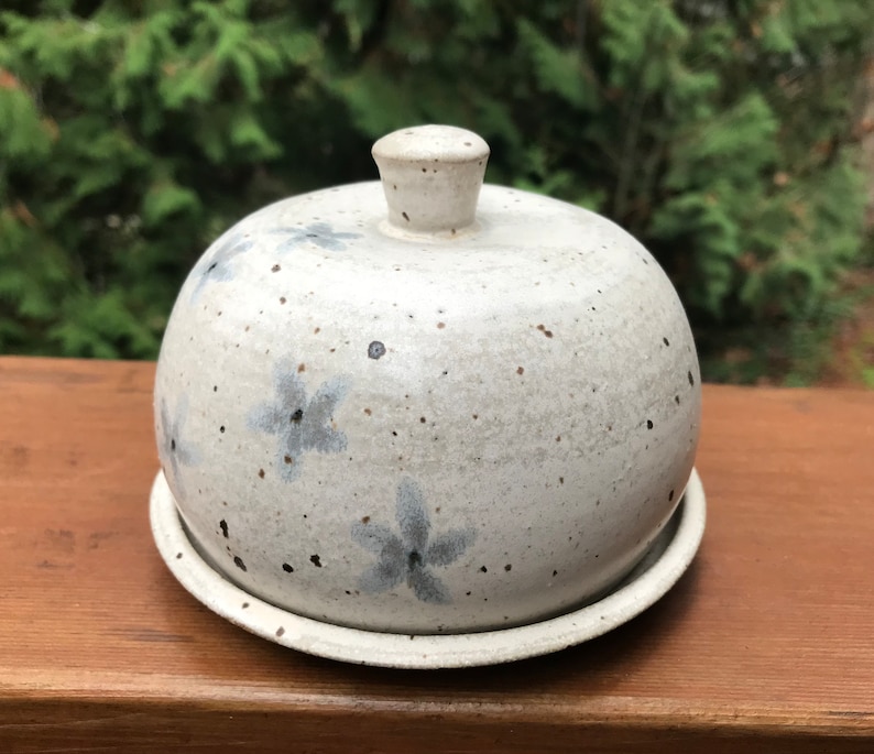 May include: White ceramic butter dish with a domed lid and a speckled glaze. The lid has a knob on top and the dish has a base. The dish is decorated with blue flower designs.