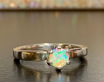 Puede incluir: Un anillo de plata con una piedra de ópalo redonda e iridiscente engastada en una montura de garras.