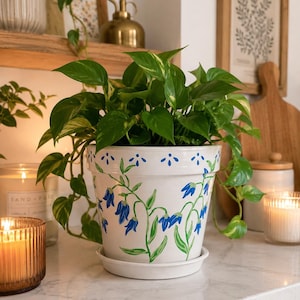 May include: A white ceramic plant pot with a matching saucer, featuring hand-painted blue bell-shaped flowers and green stems. The rim is decorated with a pattern of small blue petal motifs. A lush green pothos plant grows from the pot.